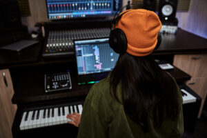 young woman, female artist looking focused while playing keyboard synthesizer, creating music, sitting in recording studio